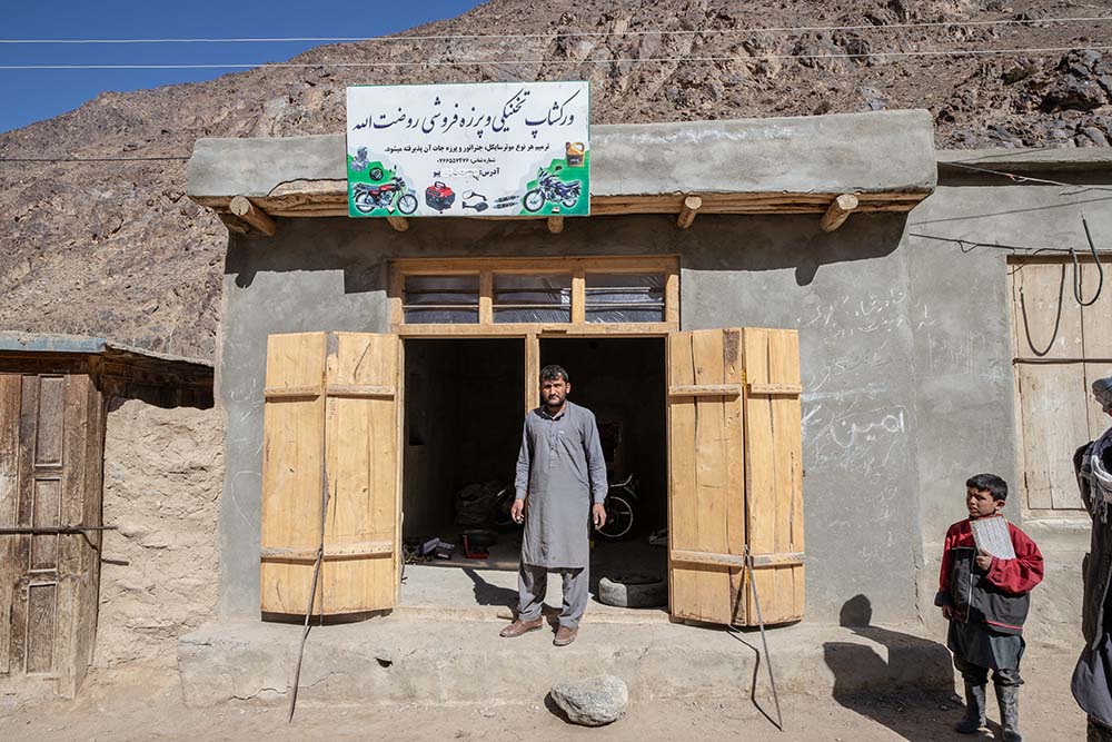 A man in Afghanistan stands in front of his mountainside motorcycle shop. The sign above him is handpainted with pictures of motorcycles and parts.