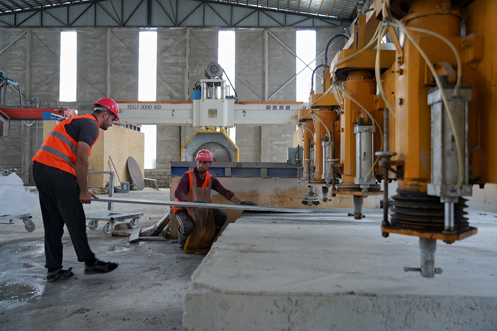 Two men in vests and hard hats operate a stone cutting machine.