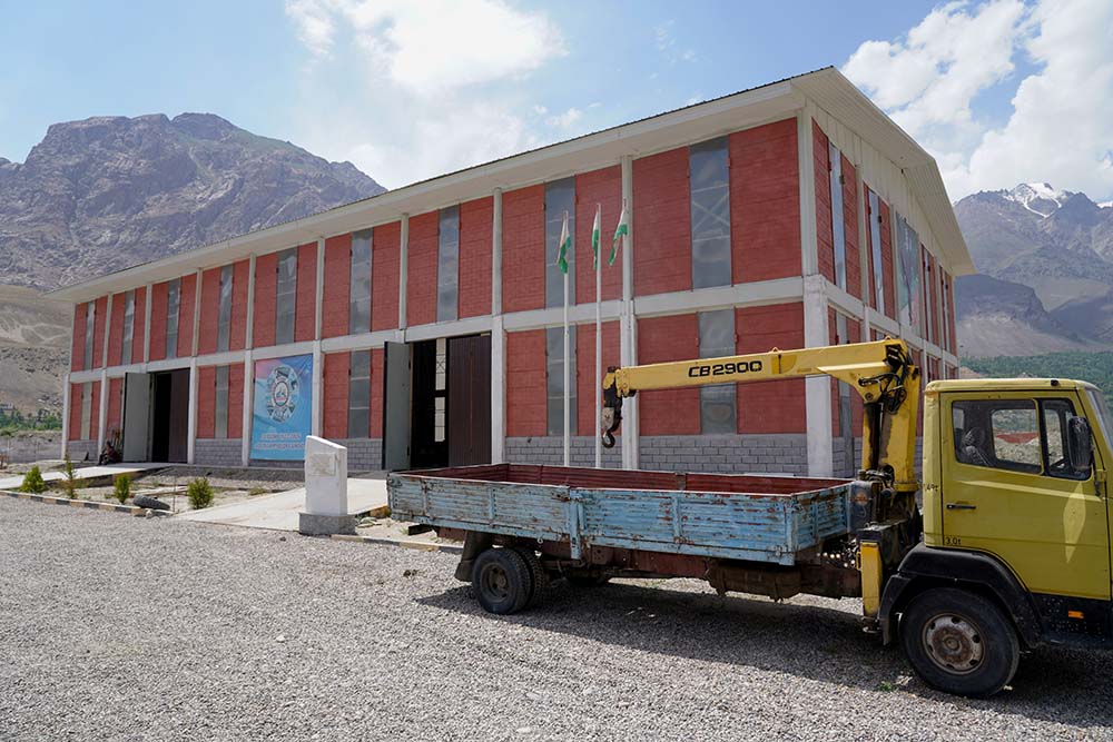 Exterior of the Pamir Stone factory, a large red brick building. A truck with crane is in the foreground, and the mountains are in the background.