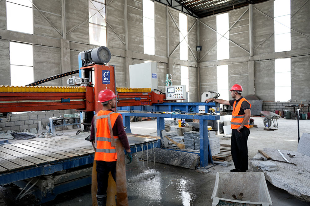 Two men in vests and hard hats operate a stone cutting machine.