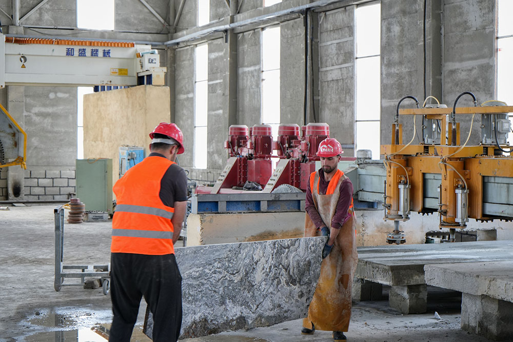 Two men in vests and hard hats carry a large slab of stone.